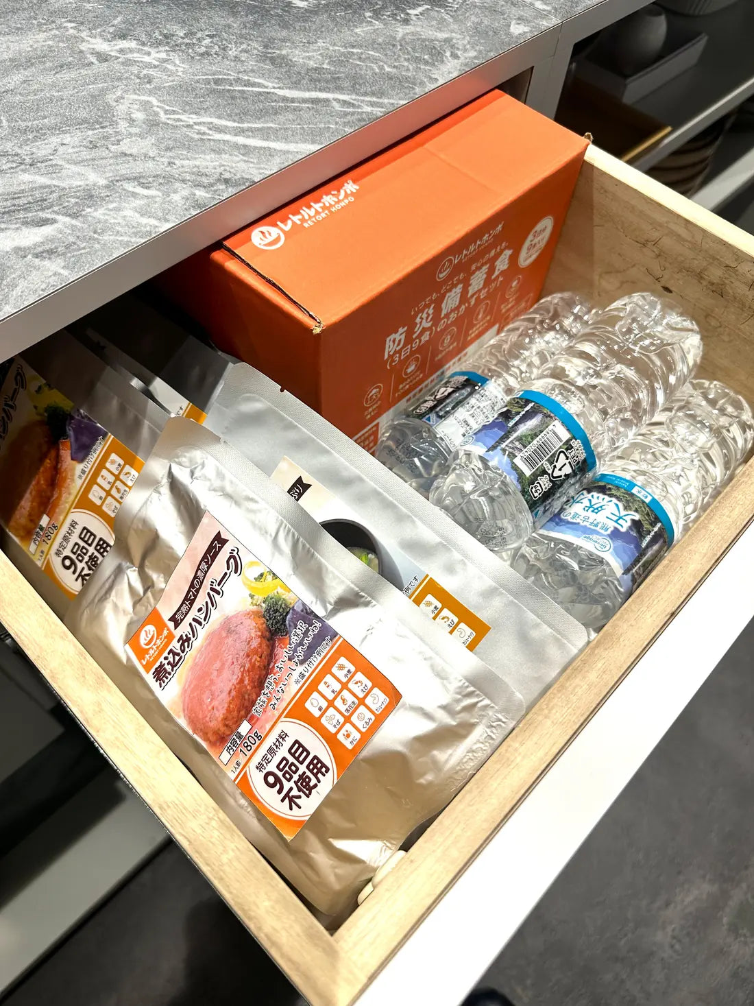 キッチンの引き出しに、レトルトホンポの防災セットの箱・煮込みハンバーグのパウチ・水のボトルを並べて備蓄しているところ